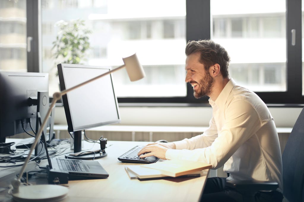 《拋棄B計畫》讀後心得:破釜沉舟開創命運的四個觀念 2 A man smiling while working at an office desk with a computer and natural daylight streaming in through large windows.