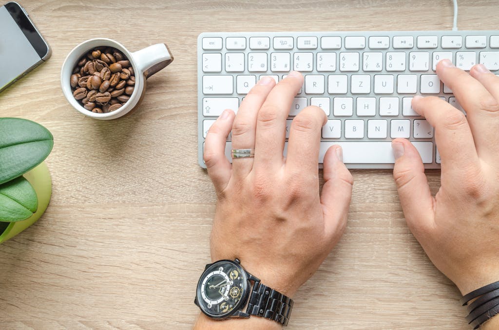 《自媒體斜槓一年,我多存了250萬!》讀後心得 3 Hands typing on a keyboard beside a cup of coffee and a smartphone on a wooden table.